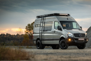 White semi-truck driving on a highway through a golden grassy valley with mountains in the background under a partly cloudy sky.